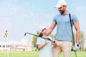 pequeno jogador de golfe com o pai. jovem feliz e seu filho segurando tacos de golfe e olhando um ao outro em pé no campo de golfe foto