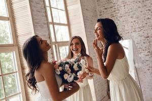 alegria sincera. mulher jovem e atraente segurando um buquê de casamento e sorrindo enquanto suas amigas bebendo champanhe foto