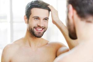 começando um novo dia com um sorriso. jovem bonito tocando seu cabelo com a mão e sorrindo em frente ao espelho foto