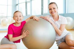 meninas no clube esportivo. alegre mãe e filha inclinando-se para a bola de fitness e sorrindo enquanto está sentado no chão no clube desportivo foto