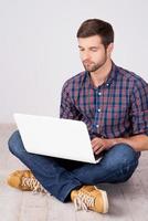 homem casual trabalhando no laptop. jovem bonito trabalhando no laptop enquanto está sentado no chão de madeira foto