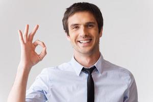 gesticulando sinal de ok. jovem alegre de camisa e gravata, gesticulando sinal de ok em pé contra um fundo cinza foto