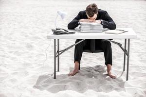 cansado do trabalho de escritório. frustrado jovem empresário inclinando a cabeça na mesa em pé na areia foto