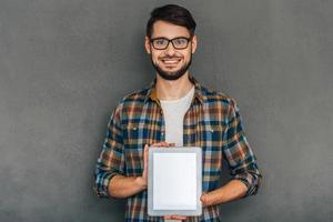 isso pode ajudá-lo jovem alegre de óculos mostrando seu tablet digital com espaço de cópia e olhando para a câmera com um sorriso em pé contra um fundo cinza foto