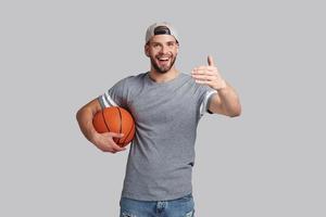 sempre pronto para vencer. bonito jovem sorridente carregando uma bola de basquete e olhando para a câmera em pé contra um fundo cinza foto
