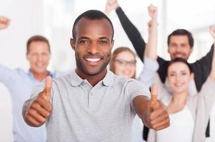 equipe de negócios feliz. feliz jovem africano mostrando seus polegares para você e sorrindo enquanto grupo de pessoas em roupas casuais em pé no fundo foto