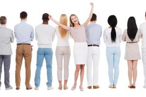 vencedor do dia-a-dia. vista traseira do grupo de pessoas em pé em uma fileira e contra um fundo branco, enquanto uma mulher em pé de frente para a câmera e expressando positividade foto