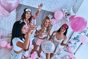jovem e linda. vista superior de quatro mulheres atraentes e sorridentes de pijama bebendo champanhe enquanto faz uma festa do pijama no quarto foto