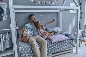 criando memórias felizes. jovem pai e sua filha tomando selfie enquanto está sentado na cama em casa foto