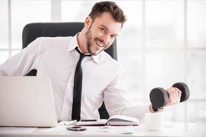 treinamento no local de trabalho. homem jovem bonito de barba de camisa e gravata treinando com haltere e sorrindo para a câmera enquanto está sentado em seu local de trabalho foto