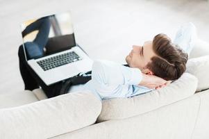 hora de fazer uma pausa. vista superior do jovem empresário segurando laptop nas pernas e segurando as mãos atrás da cabeça enquanto está sentado no sofá foto
