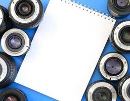 várias lentes fotográficas e caderno branco estão sobre um fundo azul brilhante. espaço para texto foto