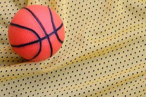 pequena bola de basquete de borracha laranja encontra-se em uma textura de tecido de roupa de camisa esportiva amarela e fundo com muitas dobras foto