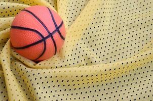 pequena bola de basquete de borracha laranja encontra-se em uma textura de tecido de roupa de camisa esportiva amarela e fundo com muitas dobras foto