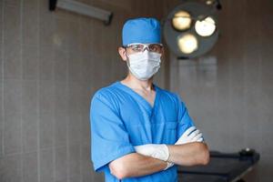 jovem médico profissional de uniforme azul, óculos e chapéu fica na sala de cirurgia. foto