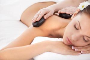 massagem com pedras quentes. bela jovem deitada na frente enquanto massagista massageando as costas com pedras de spa foto