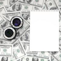 duas lentes fotográficas e um caderno branco estão no fundo de muitas notas de dólar. espaço para texto foto