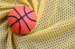 pequena bola de basquete de borracha laranja encontra-se em uma textura de tecido de roupa de camisa esportiva amarela e fundo com muitas dobras foto