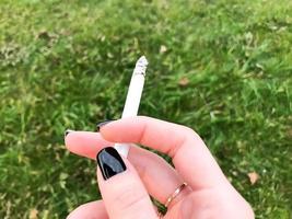 a mão de uma garota fumante, uma mulher com dedos bonitos e um anel de casamento de ouro e manicure preta nas unhas segurando um cigarro aceso no fundo da grama verde foto