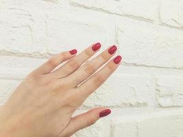 manicure brilhante, vermelho e brilhante em um fundo branco fosco. parede de tijolos de gesso para fotos de serviços de beleza de mestres. belo ensaio de manicure e maquiagem