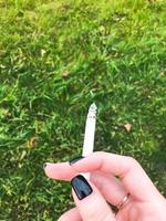 a mão de uma garota fumante, uma mulher com dedos bonitos e um anel de casamento de ouro e manicure preta nas unhas segurando um cigarro aceso no fundo da grama verde foto