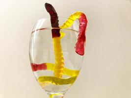 vermes gomosos flutuam em um copo transparente de água. vermes gomosos brilhantes e doces saem da água. vermes de gelatina amarelos, marrons e vermelhos dentro do vidro foto
