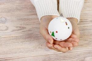 conceito de natal com mão e bola branca - brinquedo de árvore de natal. bola de natal redonda branca na mão feminina. fundo de madeira foto