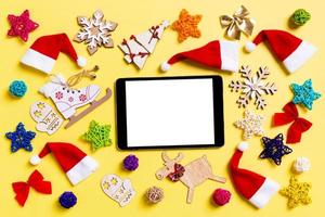 vista superior do tablet digital com enfeites de natal e chapéus de papai noel em fundo amarelo. conceito de feliz feriado foto