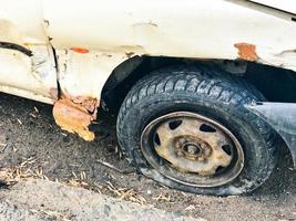 velho carro quebrado enferrujado branco da carcaça com rodas perfuradas abaixadas com corrosão de arranhões e um pára-choque rasgado com amassados e arranhões jogados na estrada. lixo automotivo foto