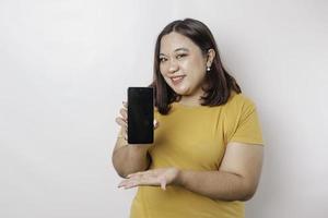 um retrato de uma mulher de tamanho grande asiática feliz está sorrindo e mostrando espaço de cópia em seu smartphone vestindo camisa amarela isolada por um fundo branco foto