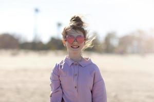 sorridente menina feliz em óculos de sol em forma de coração está se divertindo na praia. férias de verão. criança está olhando para a câmera no fundo das palmeiras foto