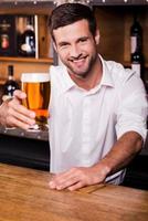 deixe-me saciar sua sede bonito jovem barman masculino de camisa branca esticando o copo com cerveja e sorrindo em pé no balcão do bar foto