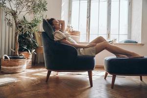 bela jovem mantendo os olhos fechados enquanto relaxa na cadeira confortável em casa foto
