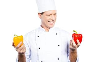 escolhendo a pimenta certa para sua refeição. alegre chef maduro em uniforme branco segurando pimentões coloridos e olhando para o vermelho em pé contra um fundo branco foto