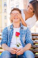 adivinhe quem bela jovem cobrindo os olhos do namorado e sorrindo enquanto o homem sentado no banco e segurando a rosa vermelha foto