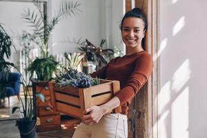jovem alegre carregando caixa de madeira com plantas enquanto se inclinava na porta em casa