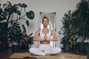 bela jovem meditando enquanto está sentado no chão com plantas de casa ao seu redor foto