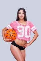 eu amo basquete atraente jovem líder de torcida feminina segurando uma bola de basquete e olhando para a câmera em pé contra um fundo cinza foto