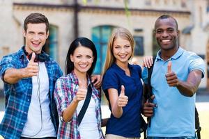 adoramos estudar quatro jovens felizes mostrando os polegares para cima e sorrindo enquanto estamos perto um do outro ao ar livre foto