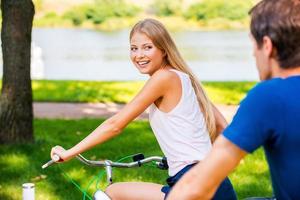 estou ganhando linda jovem sorridente andando de bicicleta e olhando por cima do ombro enquanto o namorado andando atrás dela foto