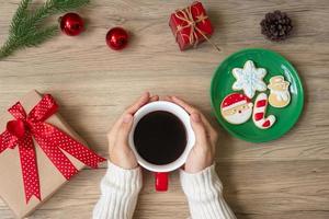 feliz natal com mão de mulher segurando a xícara de café e biscoito caseiro na mesa. véspera de natal, festa, feriado e feliz ano novo conceito foto