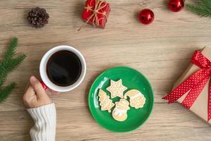 feliz natal com mão de mulher segurando a xícara de café e biscoito caseiro na mesa. véspera de natal, festa, feriado e feliz ano novo conceito foto