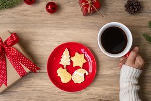 feliz natal com mão de mulher segurando a xícara de café e biscoito caseiro na mesa. véspera de natal, festa, feriado e feliz ano novo conceito foto