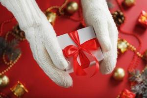 mãos em luvas de malha brancas segurando um presente em um fundo vermelho. foto