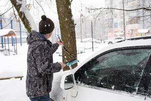 um homem escova a neve de um carro após uma nevasca. segurança rodoviária, condições meteorológicas difíceis no inverno foto