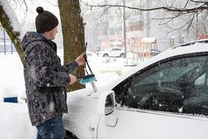 um homem escova a neve de um carro após uma nevasca. segurança rodoviária, condições meteorológicas difíceis no inverno foto