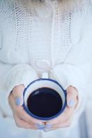 vista superior e feche as mãos da mulher segurando uma xícara quente de café ou chá em fundo de clima frio, vestindo roupas de malha de pele quente foto