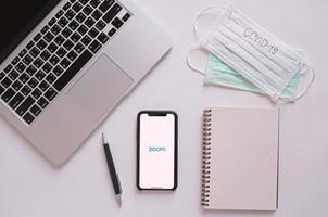 postura plana da mesa do espaço de trabalho com smartphone mostrando aplicativo de zoom e laptop com máscara e texto covid-19. zoom é uma reunião popular na nuvem ou videoconferência durante a pandemia de covid-19 e coronavírus foto