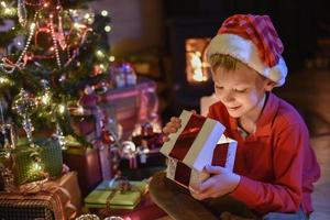 época de natal, garotinho abrindo um presente perto de uma árvore foto
