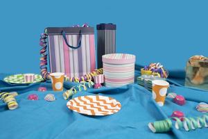 Mesa bagunça com sacolas e caixas de presente, placas de plástico e xícaras em uma mesa com uma toalha de mesa azul contra um fundo azul claro foto